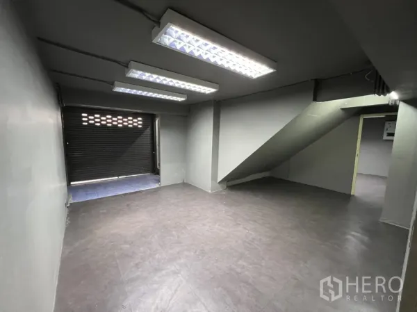 Shophouse for rent in Phra Khanong, Bangkok - Back room with staircase access, fluorescent lighting and a roller shutter door suitable for loading.