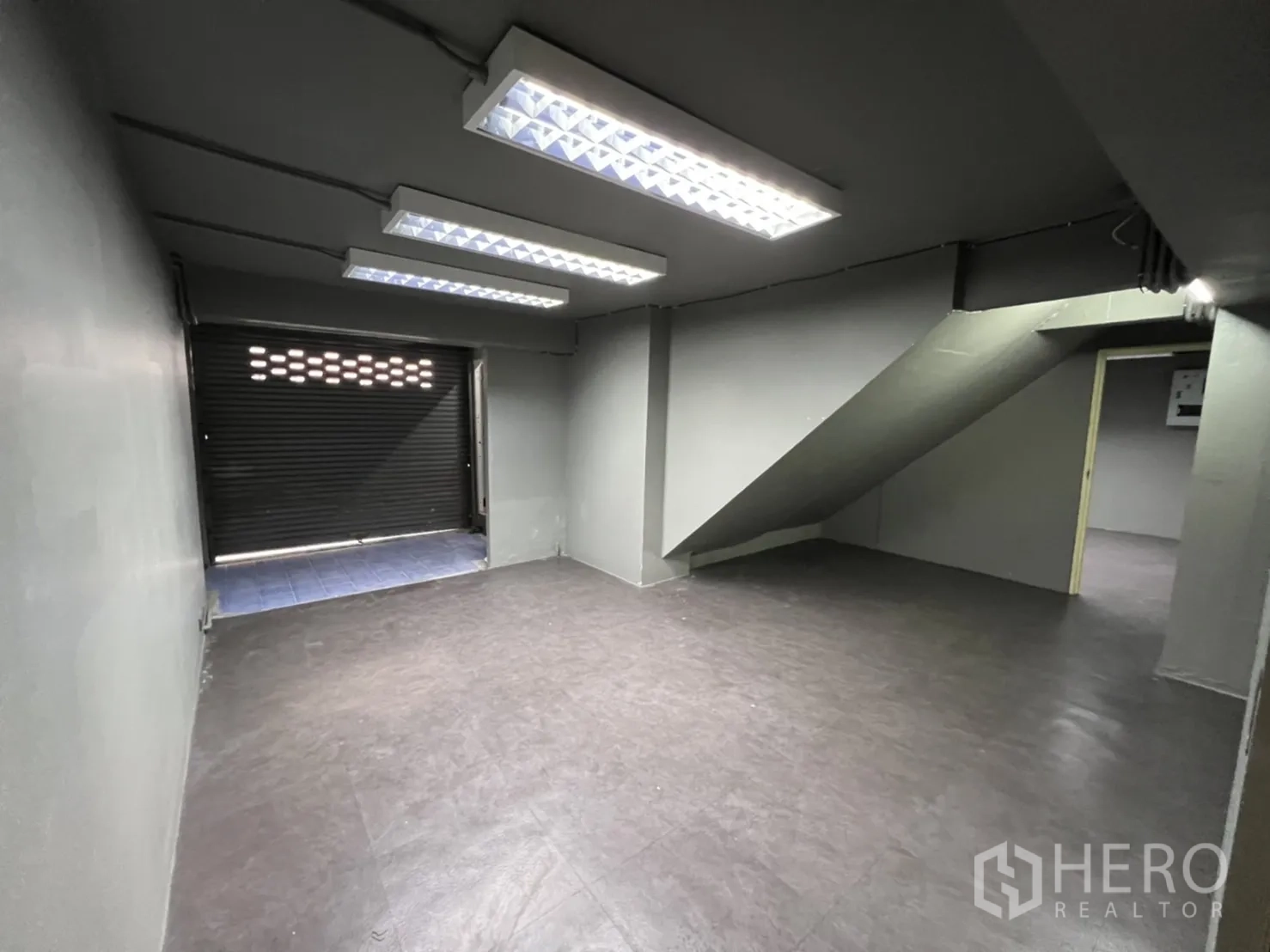 Shophouse for rent in Phra Khanong, Bangkok - Back room with staircase access, fluorescent lighting and a roller shutter door suitable for loading.
