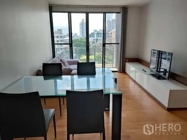 Condominium for sale in Khlong Toei, Bangkok - Living and dining room with glass table, sofa, TV console and floor-to-ceiling sliding doors to a balcony with city view.