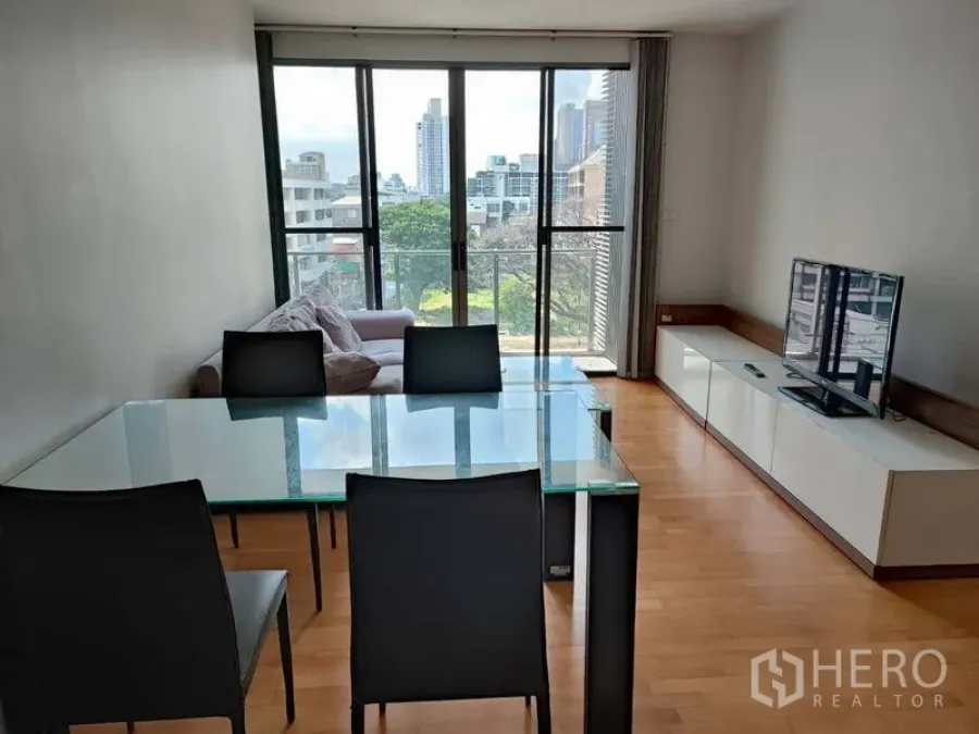 Condominium for sale in Khlong Toei, Bangkok - Living and dining room with glass table, sofa, TV console and floor-to-ceiling sliding doors to a balcony with city view.