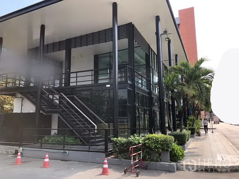 Retail Space for rent in Khlong Toei, Bangkok - Side view of retail unit with black steel frame, external staircase, large windows and sidewalk greenery.