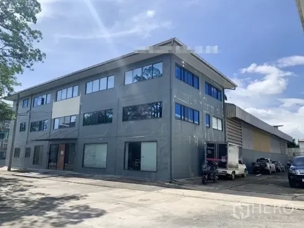 Warehouse for rent in Saphan Sung, Bangkok - Three-storey office building attached to a warehouse with parking and loading area under a clear sky.