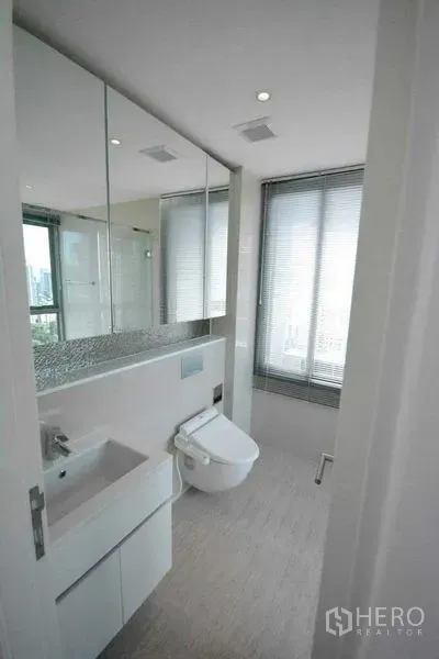 Condominium for rent or sale in Phra Khanong, Bangkok - Modern bathroom with mirrored cabinets, vanity sink and bidet toilet by a window.