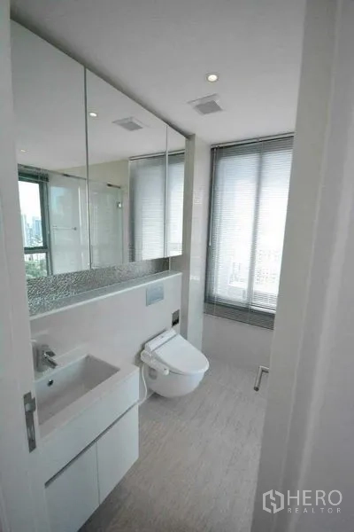 Condominium for rent or sale in Phra Khanong, Bangkok - Modern bathroom with mirrored cabinets, vanity sink and bidet toilet by a window.