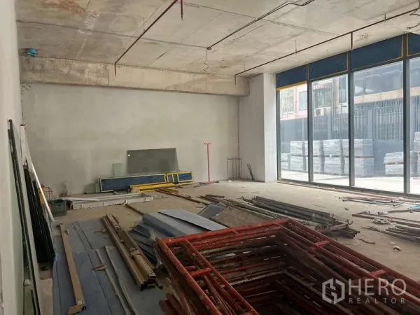 Retail Space for rent - Bare-shell ground-floor retail unit with high concrete ceiling, floor-to-ceiling glass frontage and construction materials stacked inside.
