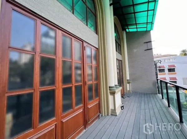 Shophouse for rent in Watthana, Bangkok - Upper-level balcony of the Sukhumvit shophouse with glass railing, arched columns, and wood-framed doors beneath a green canopy.