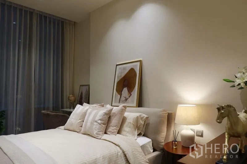 Condominium for rent in Pathum Wan, Bangkok - Serene bedroom with upholstered bed, soft neutral bedding, bedside lamp and tall curtains.