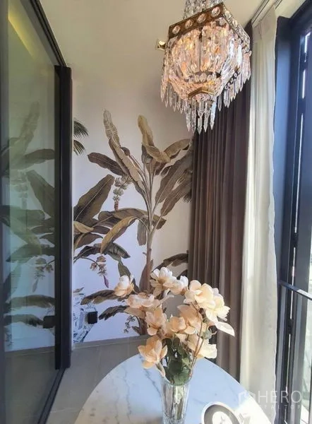 Condominium for rent in Khlong Toei, Bangkok - Close‑up of the sunlit nook with crystal pendant, floral wallpaper, round table and vase by tall windows.