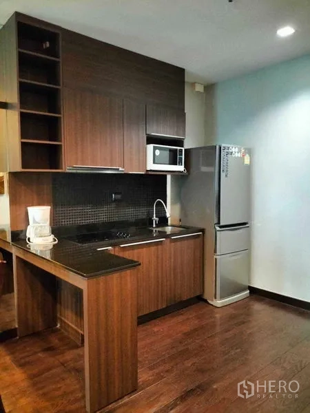 Condominium for rent in Ratchathewi, Bangkok - Compact kitchen with wood cabinetry, black countertop, microwave and fridge.