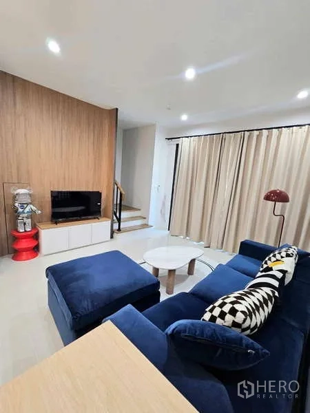Townhouse for rent in Suan Luang, Bangkok - Living room with blue sofa, built-in TV cabinet, wooden feature wall and staircase.