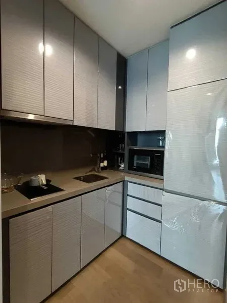 Condominium for rent or sale in Bang Rak, Bangkok - Compact L-shaped kitchen with sleek cabinetry, cooktop, sink, microwave and full-size fridge.