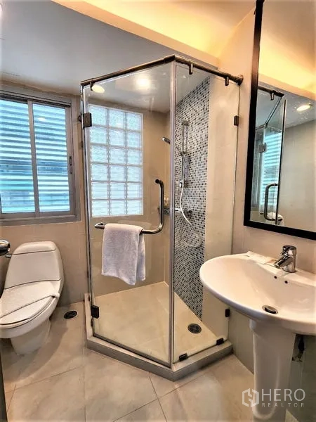 Condominium for rent in Bang Rak, Bangkok - Compact bathroom with glass shower cabin, toilet, pedestal sink and mosaic tile accent wall.