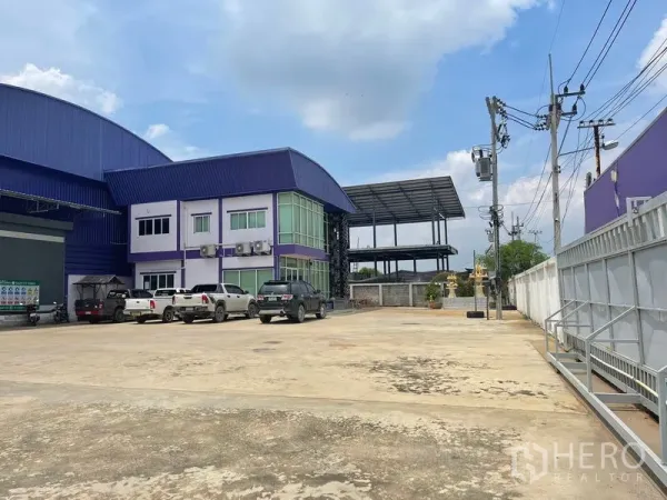 Warehouse for sale in Lam Luk Ka, Pathum Thani - Concrete parking yard beside the two-storey office building with green-tinted windows and a steel-framed covered area to the rear.