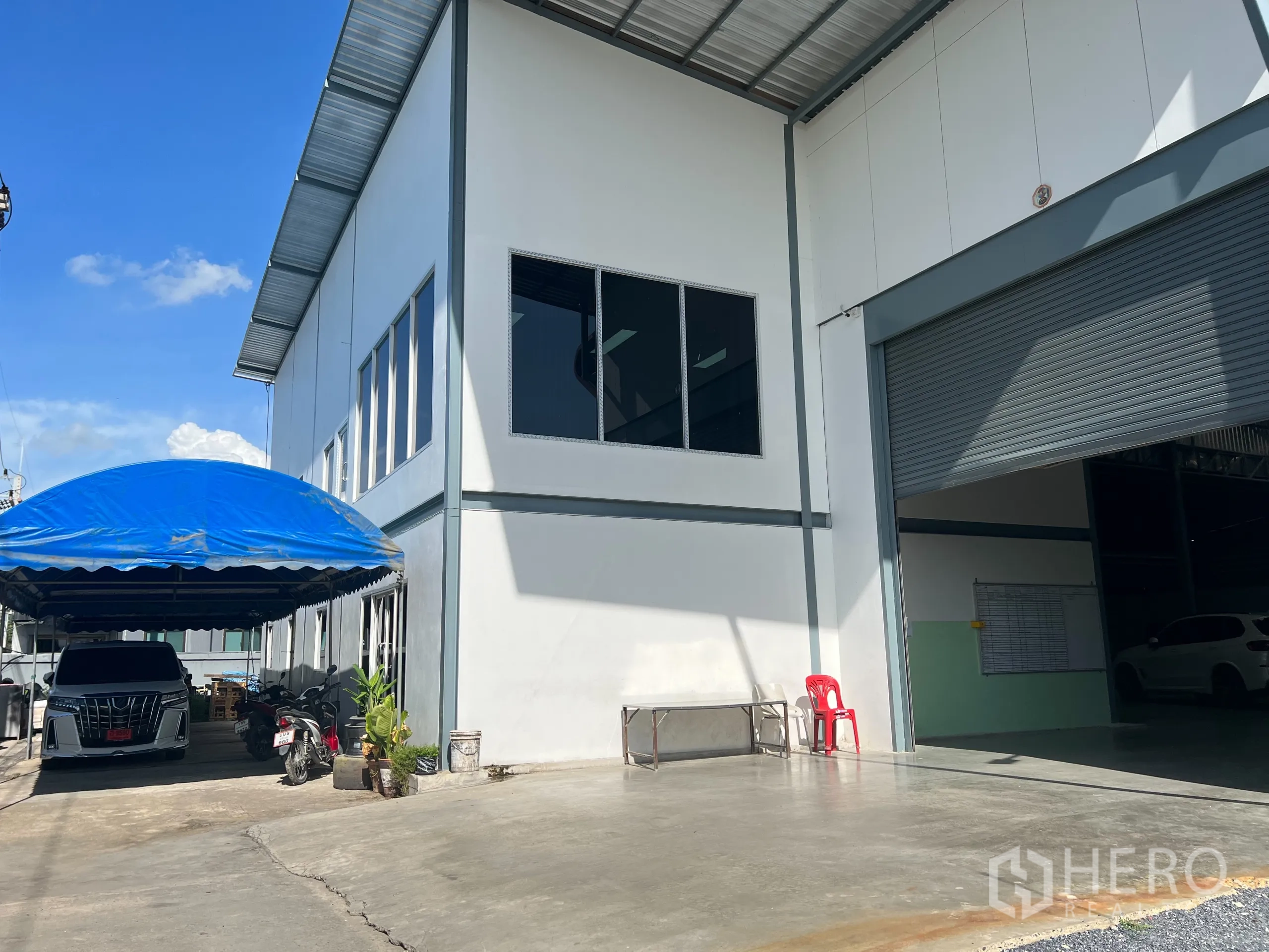 Warehouse for sale in Lam Luk Ka, Pathum Thani - Front yard showing blue carport, office windows, and a large roller shutter opening.