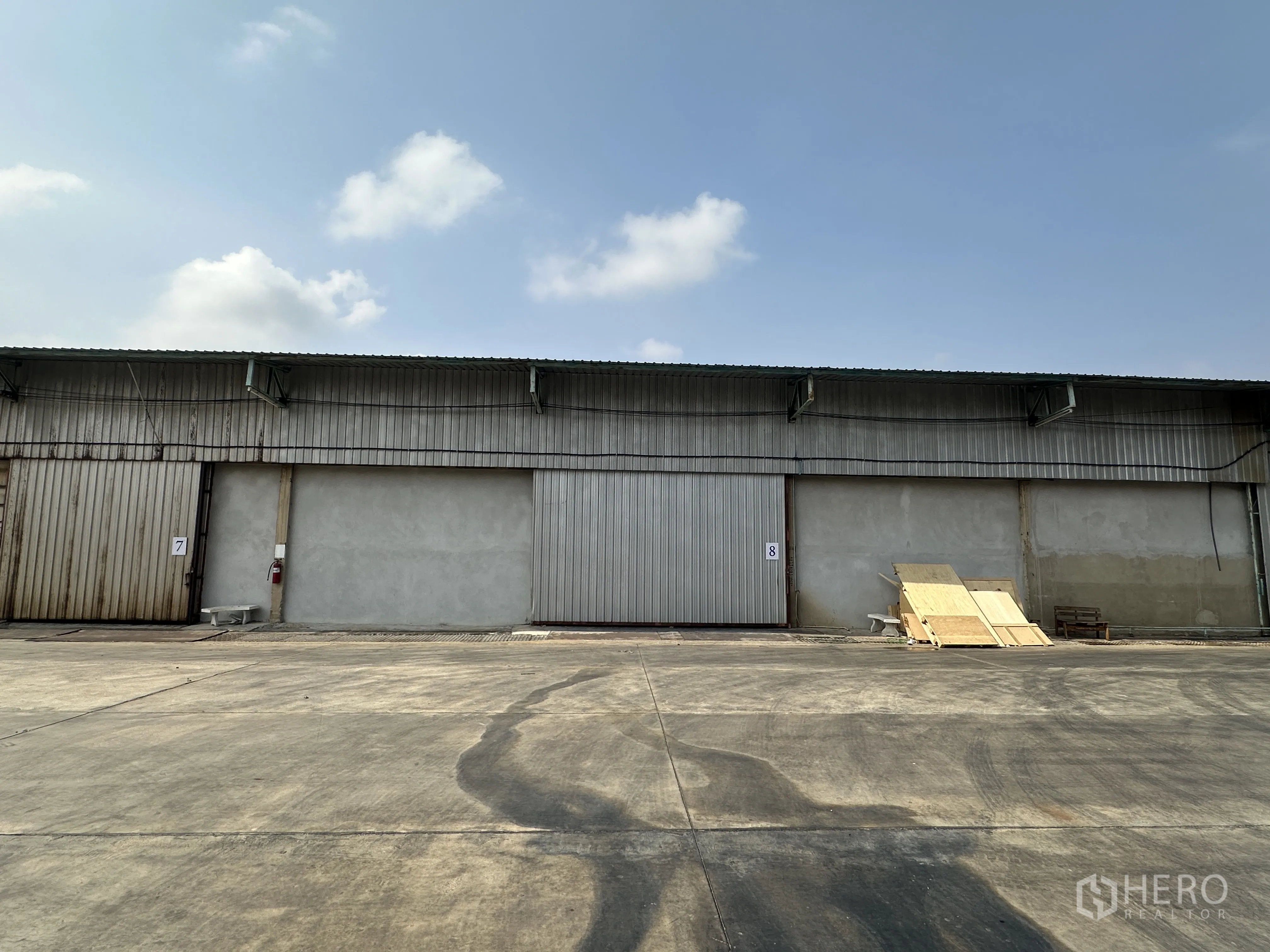 Warehouse for rent in Mueang Pathum Thani, Pathum Thani - Front exterior of a metal-clad warehouse with large sliding doors and a wide concrete yard under a clear sky.