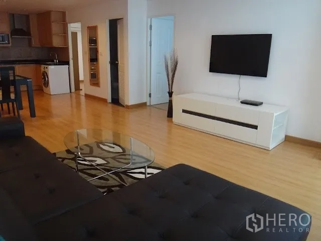 Condominium for rent in Bang Rak, Bangkok - Modern living room with a black leather sofa, glass coffee table, area rug, and wall-mounted TV in an open-plan layout.