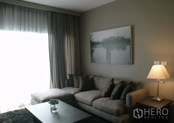 คอนโด ให้เช่าในWatthana, Bangkok - ห้องนั่งเล่นโทนเทา โซฟาตัวแอล ม่านชั้นคู่ ภาพแขวน และโคมไฟตั้งโต๊ะ