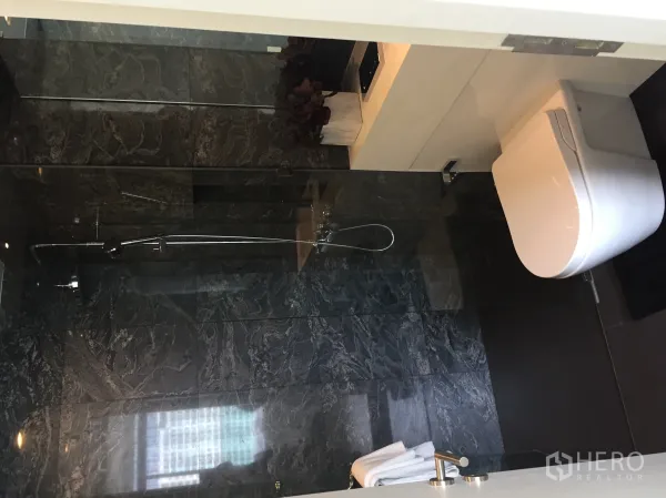 Condominium for sale in Bang Rak, Bangkok - Modern bathroom with dark stone tiles, glass shower enclosure and wall-hung toilet.