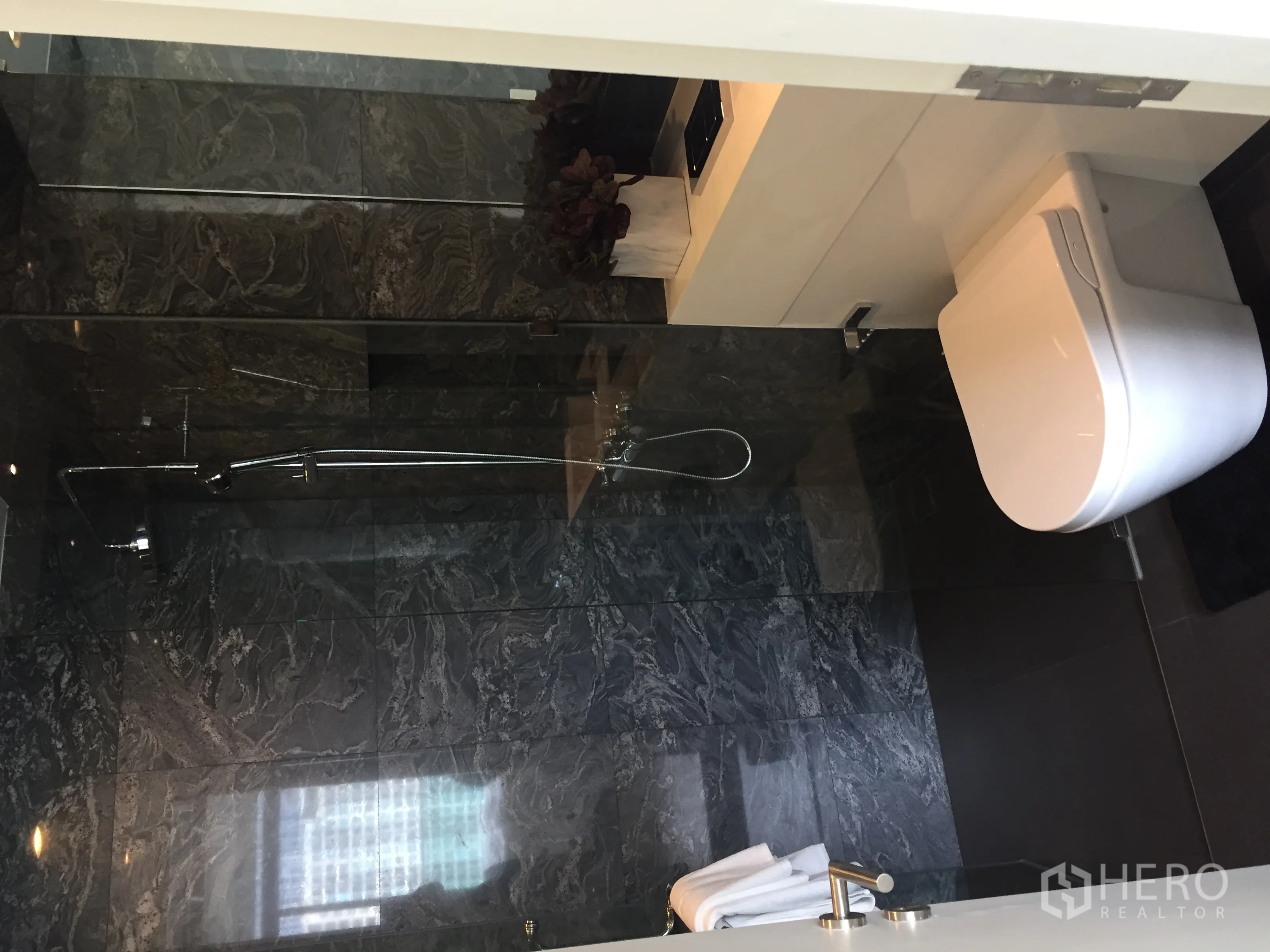 Condominium for sale in Bang Rak, Bangkok - Modern bathroom with dark stone tiles, glass shower enclosure and wall-hung toilet.