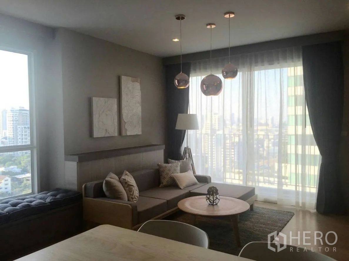 公寓 出租在Watthana, Bangkok - 明亮客厅，转角沙发、圆形茶几、吊灯及落地窗，远眺城市景观。