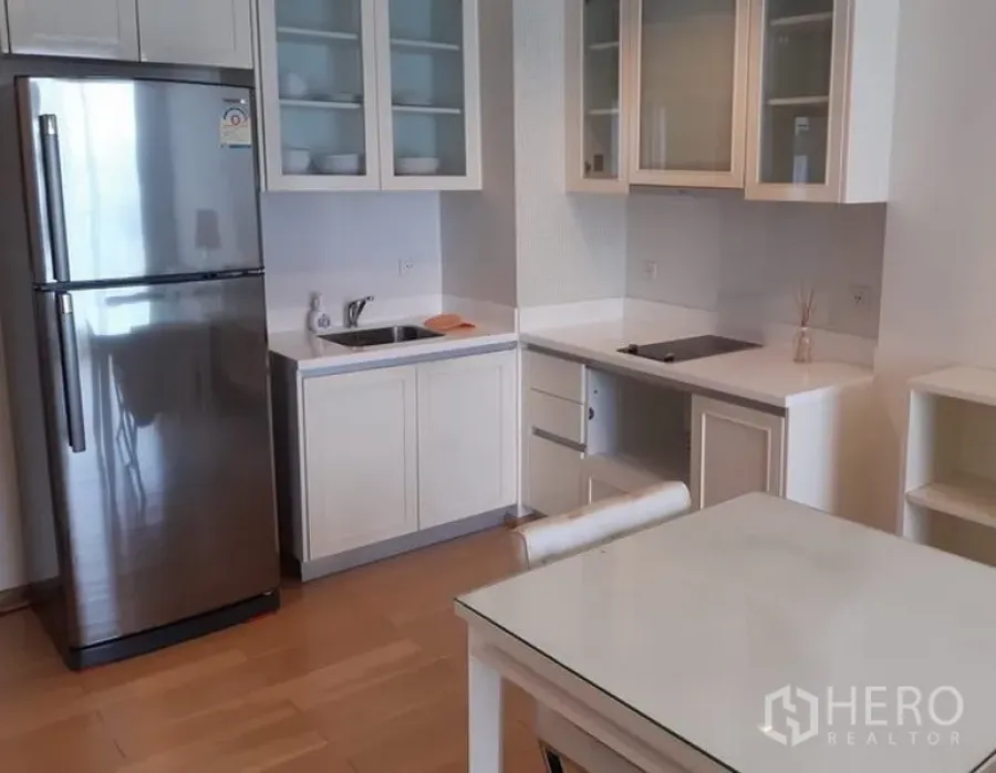 Condominium for rent in Khlong Toei, Bangkok - Bright compact kitchen with white cabinets, glass-front cupboards, stainless refrigerator, induction hob and dining table on wood flooring.