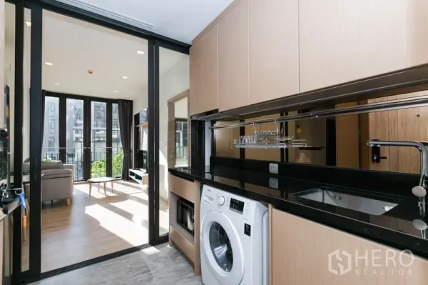 Condominium for rent in Watthana, Bangkok - Galley kitchen with black granite countertop, upper cabinets, sink and in-unit washing machine behind a glass door.