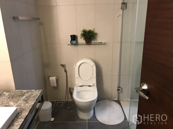 Condominium for rent or sale in Bang Rak, Bangkok - Bright bathroom with toilet, glass shower, vanity, and handheld bidet spray.