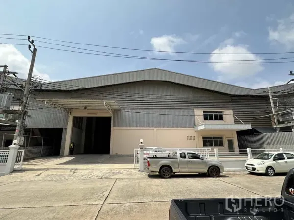Factory for rent in Samut Prakan - Front elevation of a large industrial factory with wide loading entrance, gated frontage and parking on a concrete road.