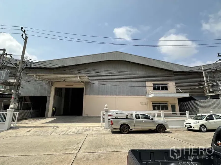 Factory for rent in Samut Prakan - Front elevation of a large industrial factory with wide loading entrance, gated frontage and parking on a concrete road.