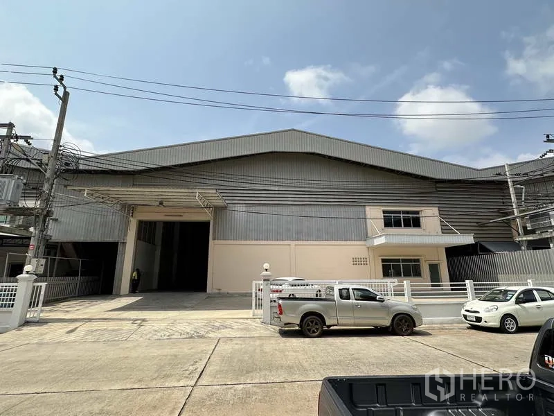 Factory for rent in Samut Prakan - Front elevation of a large industrial factory with wide loading entrance, gated frontage and parking on a concrete road.
