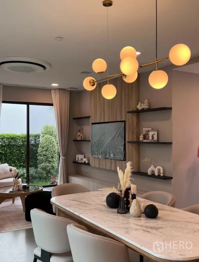 Detached House for rent in Samut Prakan - Elegant dining and living area with a marble table, globe pendant light, built-in TV wall and garden view through sliding doors.