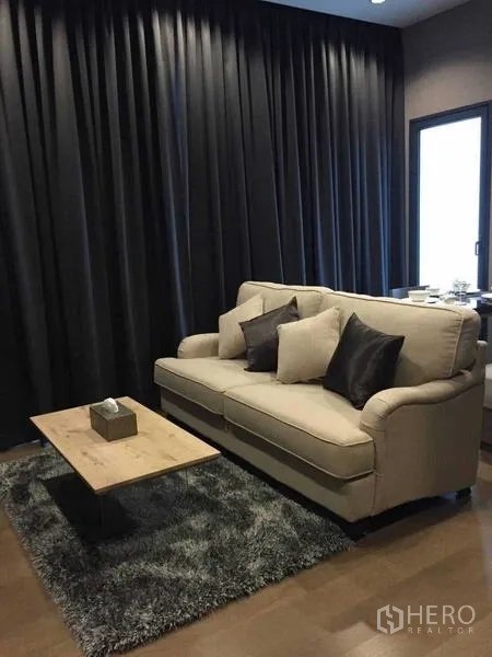 Condominium for rent in Bang Rak, Bangkok - Living area with beige sofa, wooden coffee table, and dark drapes on wood floors.