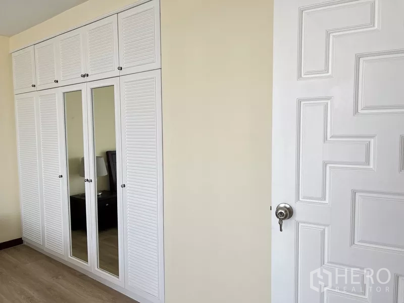 Condominium for rent in Khlong Toei, Bangkok - Bedroom wall fitted with tall white built-in wardrobes and mirrored doors.