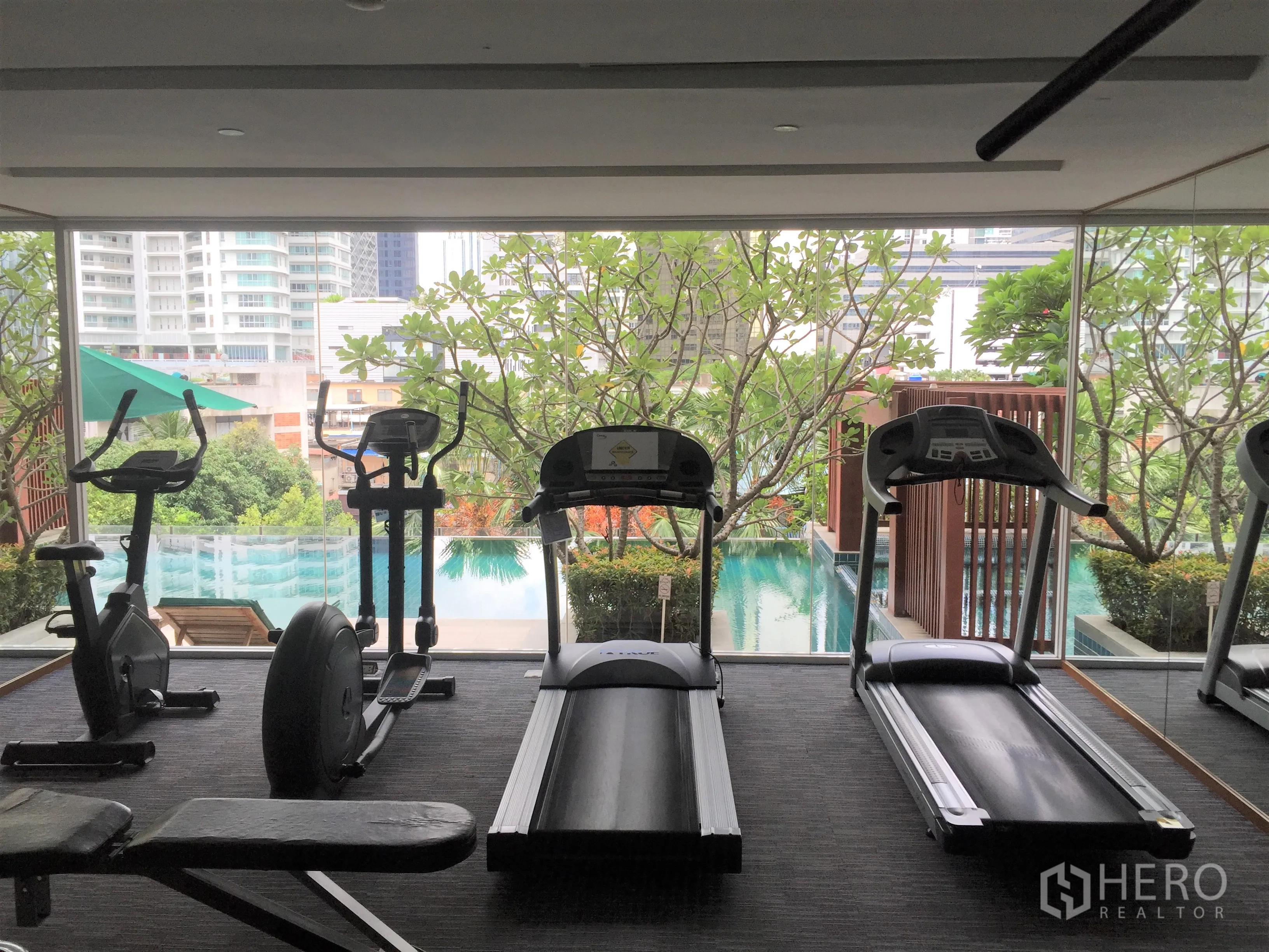 公寓 出租或出售在Watthana, Bangkok - 健身房内跑步机与器械，窗外为景观泳池与绿化。