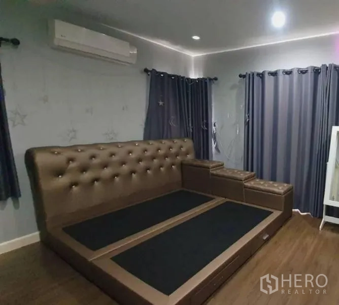 独立式住宅 出售在Pathum Thani - 温馨卧室，纽扣软包床架、深色窗帘与木地板。