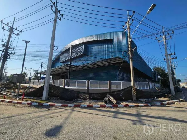 โรงงาน ให้เช่าในChon Buri - มุมอาคารโรงงานผนังเมทัลชีทสีน้ำเงิน พร้อมรั้วรอบและเสาไฟฟ้าภายนอก