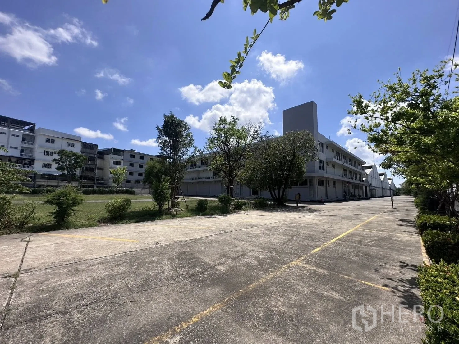 Warehouse for rent in Mueang Samut Prakan, Samut Prakan - Large concrete yard with parking lines, trees, and a low-rise industrial building.