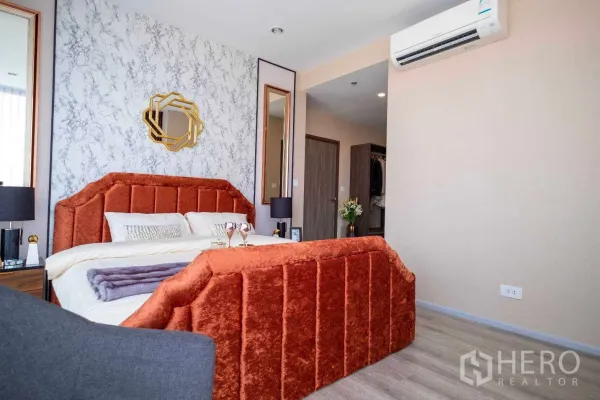 公寓 出租在Bang Na, Bangkok - 卧室角度展示橘色软包床、石纹墙、双面镜与空调，连通更衣区。