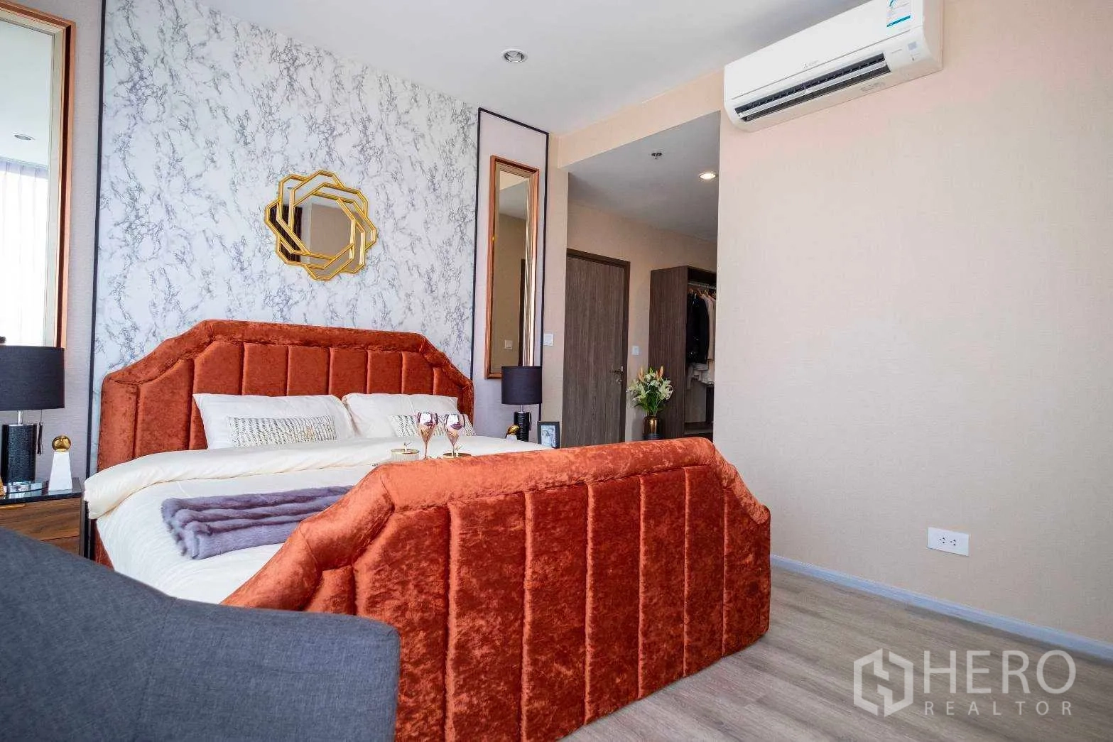 公寓 出租在Bang Na, Bangkok - 卧室角度展示橘色软包床、石纹墙、双面镜与空调，连通更衣区。