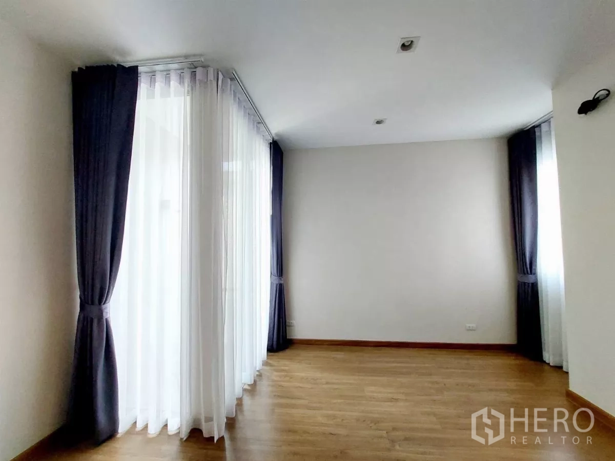 Townhouse for rent in Lat Phrao, Bangkok - Bright corner bedroom with wood flooring and floor-to-ceiling windows dressed in sheer and blackout curtains.