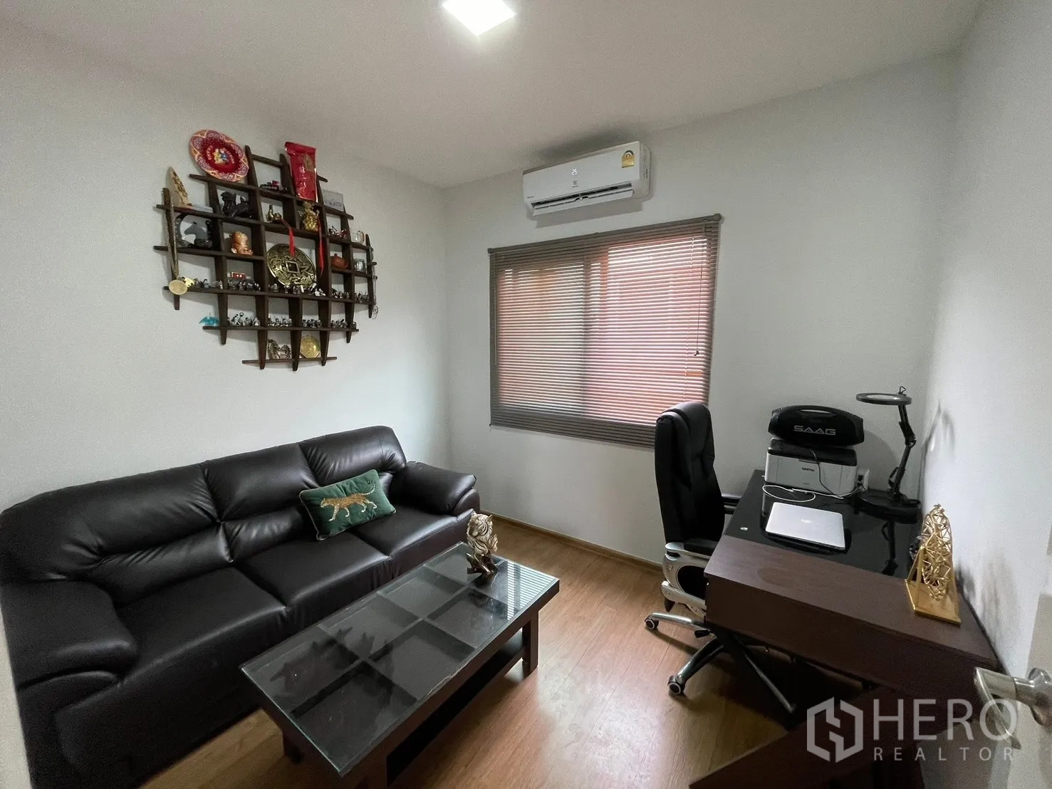ทาวน์เฮาส์ for rent in Bang Phli, Samut Prakan - ห้องนั่งเล่นทำงานกะทัดรัด โซฟาหนังสีดำ โต๊ะกระจก ชั้นวางของติดผนัง และโต๊ะทำงานใต้หน้าต่างมู่ลี่ พร้อมเครื่องปรับอากาศ