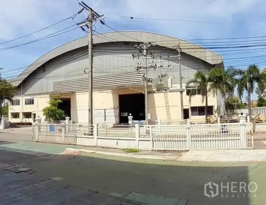 โรงงาน ให้เช่าในMueang Samut Prakan, Samut Prakan - มุมมองด้านหน้าโรงงานหลังคาโค้งขนาดใหญ่ พร้อมรั้วหน้า ปาล์ม และสายไฟเหนือถนน