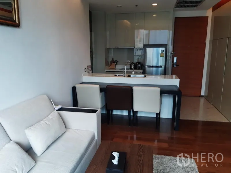 Condominium for rent in Khlong Toei, Bangkok - Compact modern kitchen with island breakfast bar, built-in cabinets and stainless refrigerator beside a cozy living area.