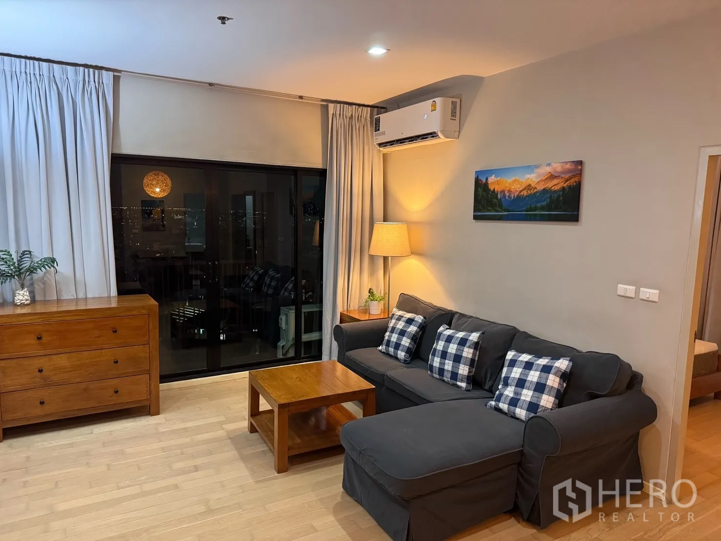 คอนโด for rent or sale in Watthana, Bangkok - ห้องนั่งเล่นอบอุ่นด้วยโซฟาสีเข้ม โต๊ะไม้ เครื่องปรับอากาศ และประตูกระจกออกสู่ระเบียงวิวเมืองยามค่ำคืน