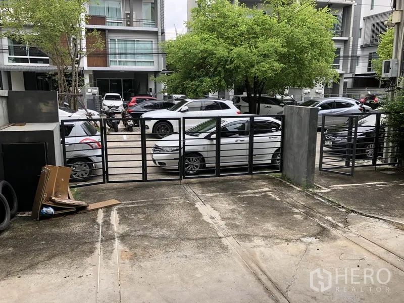 带办公室的家 出租在Wang Thonglang, Bangkok - 前院带铁门的停车区，面向绿荫街道与多辆停放车辆。
