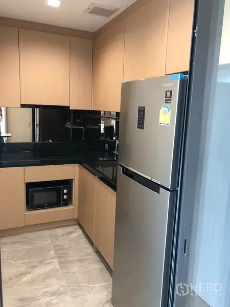 Condominium for rent in Watthana, Bangkok - Compact U-shaped kitchen with wood cabinets, black countertop, glass backsplash, microwave and stainless refrigerator.