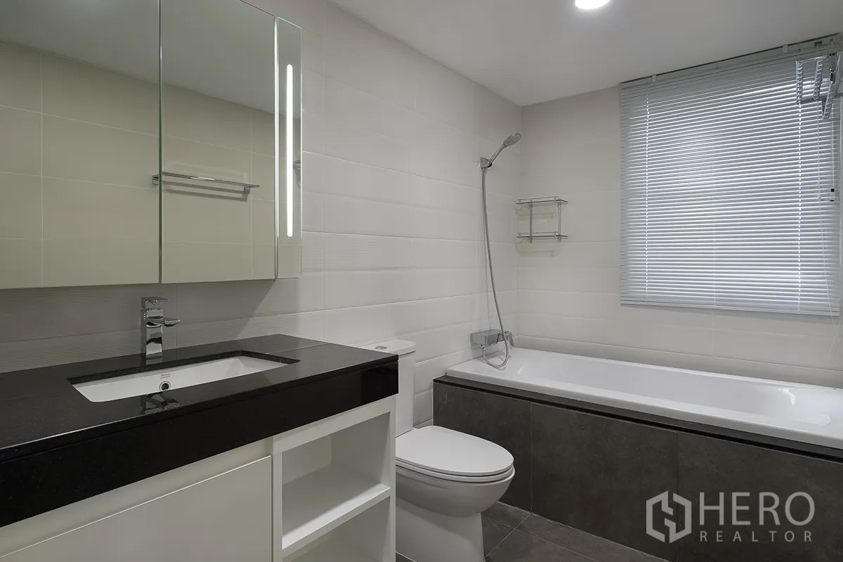Condominium for rent in Khlong Toei, Bangkok - Bright bathroom with bathtub and handheld shower, mirrored cabinet, and window blinds.