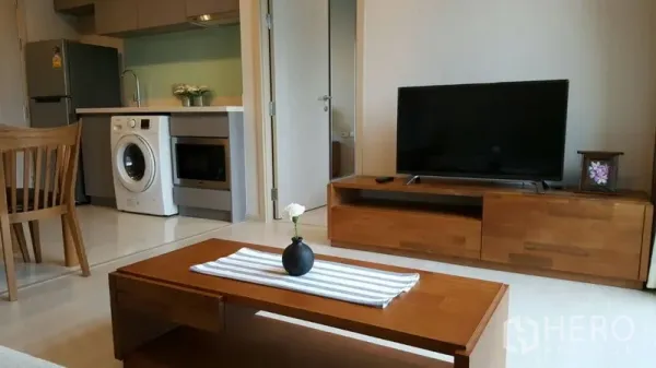 Condominium for rent in Khlong Toei, Bangkok - Living area with TV console, open-plan kitchen and washing machine in view.