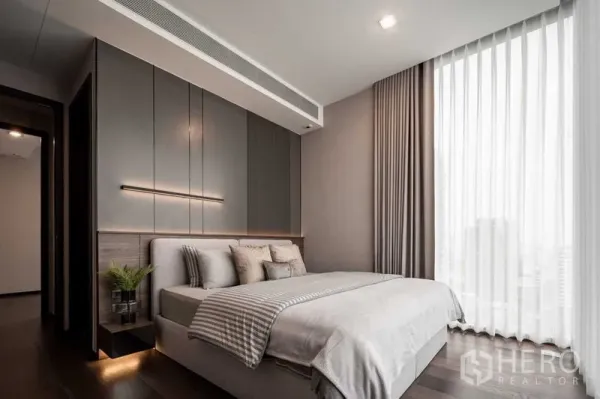 Condominium for rent in Watthana, Bangkok - Modern master bedroom with upholstered bed, grey panel wall and large window with sheer drapes.
