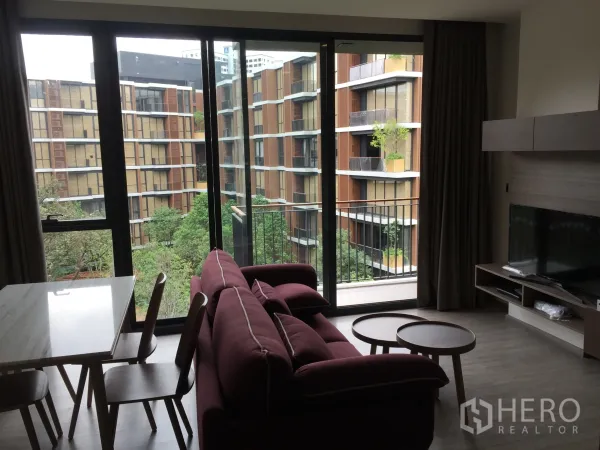 คอนโด ให้เช่าในWatthana, Bangkok - ห้องนั่งเล่นโซฟาสีแดงไวน์และทีวี เปิดสู่ระเบียงผ่านหน้าต่างสูงจรดเพดาน มองเห็นสวนส่วนกลางร่มรื่น.